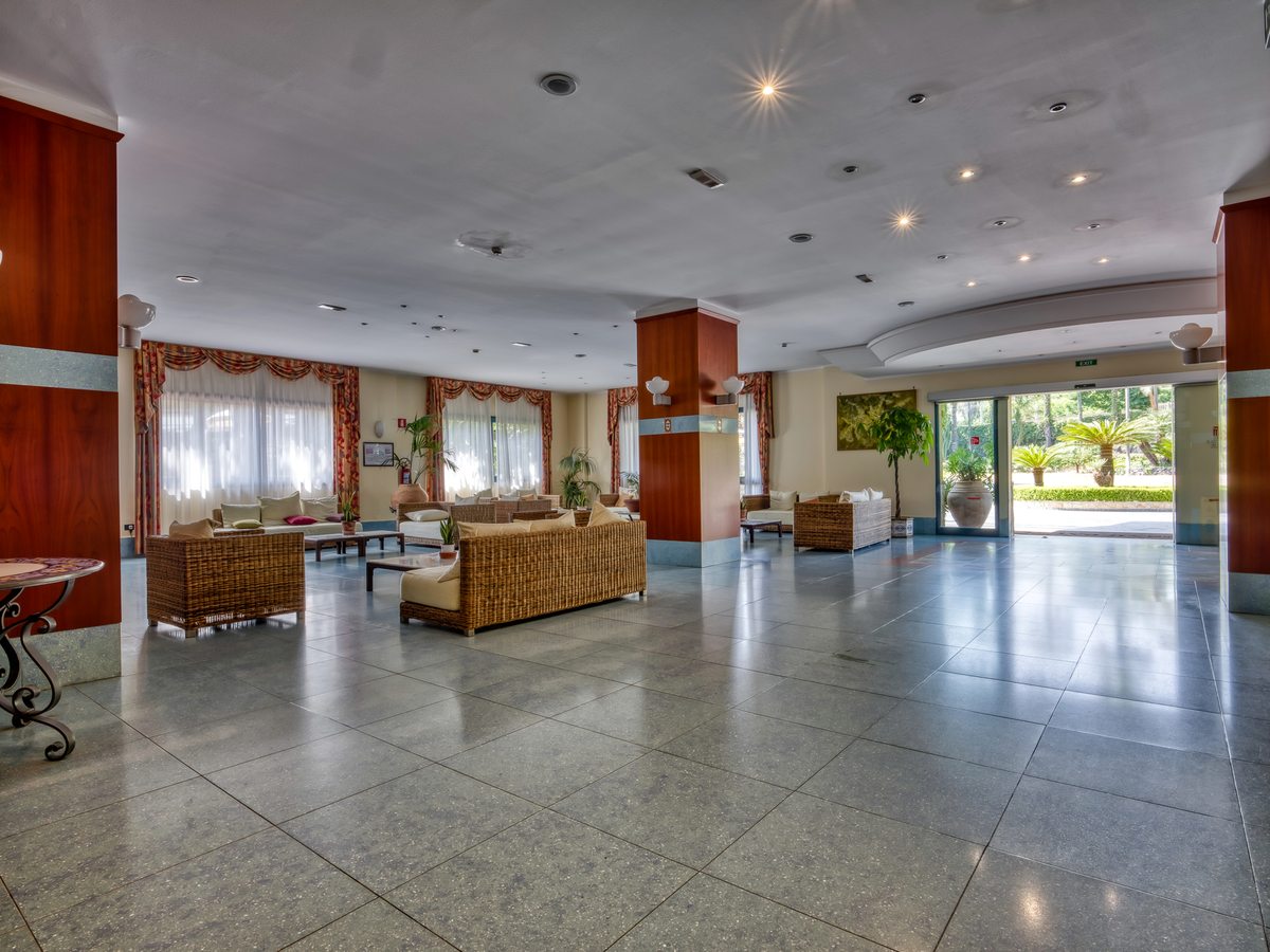 Open-plan hotel lobby with woven rattan sofas and low tables on a polished grey marble floor, curtained windows to one side and glass doors opening to a garden.