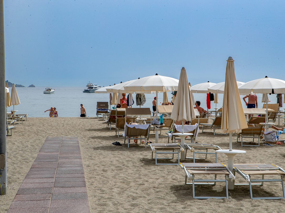 A sandy beach with rows of cream parasols and sun loungers, a paved walkway leading to a calm sea with moored boats and a distant headland.