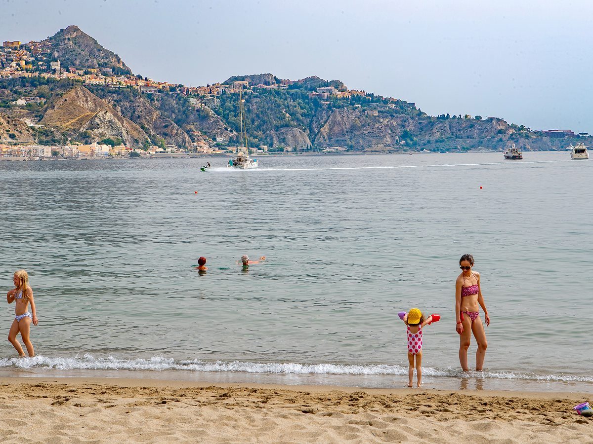 A mother and toddler paddling at the edge of a calm sea, with swimmers beyond and the hilltop town of Taormina on the headland in the distance.
