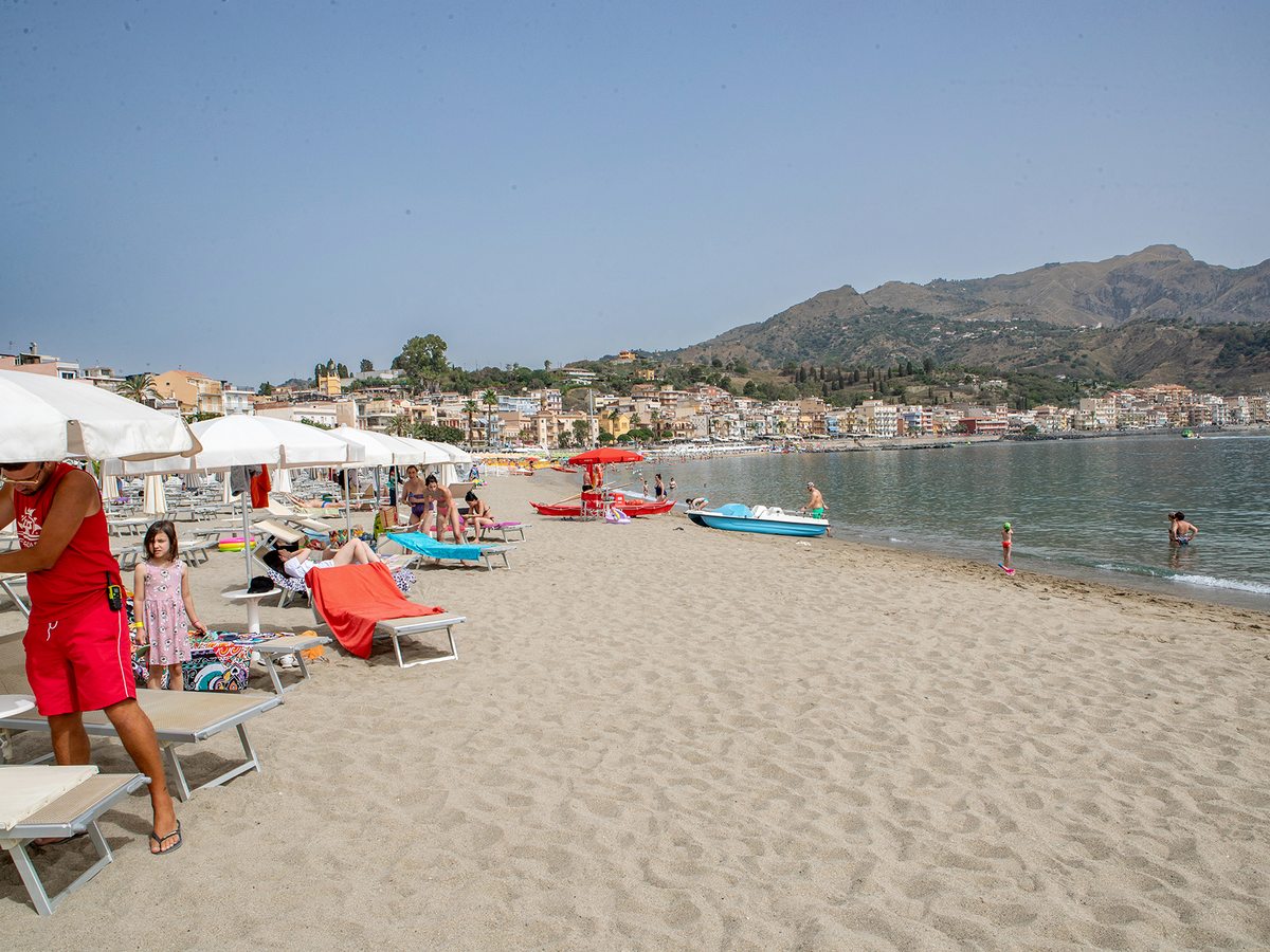 A long sandy beach with parasols and pedalos, the town of Giardini Naxos stretching along the shoreline and mountains rising behind.