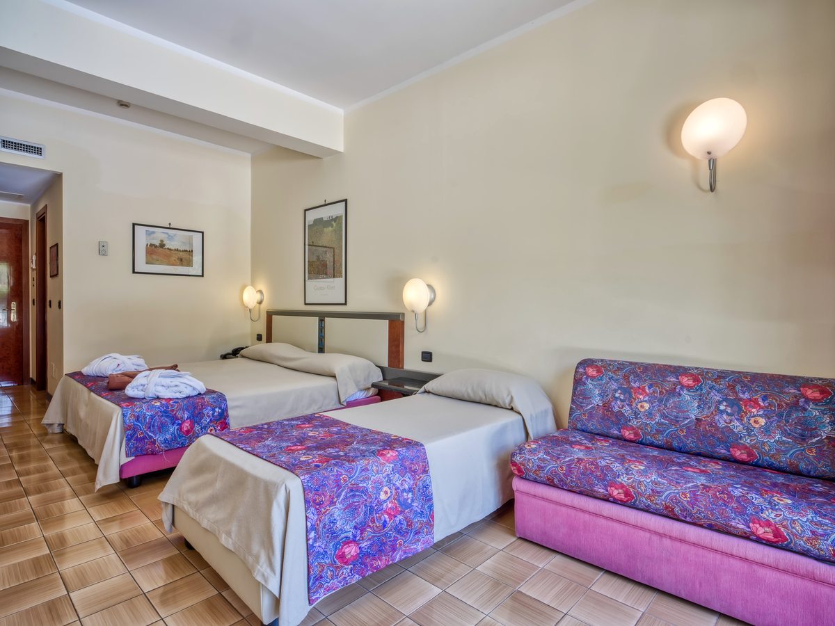 A family room with two beds and a pink floral sofa bed against the wall, framed landscape prints and a tiled floor.