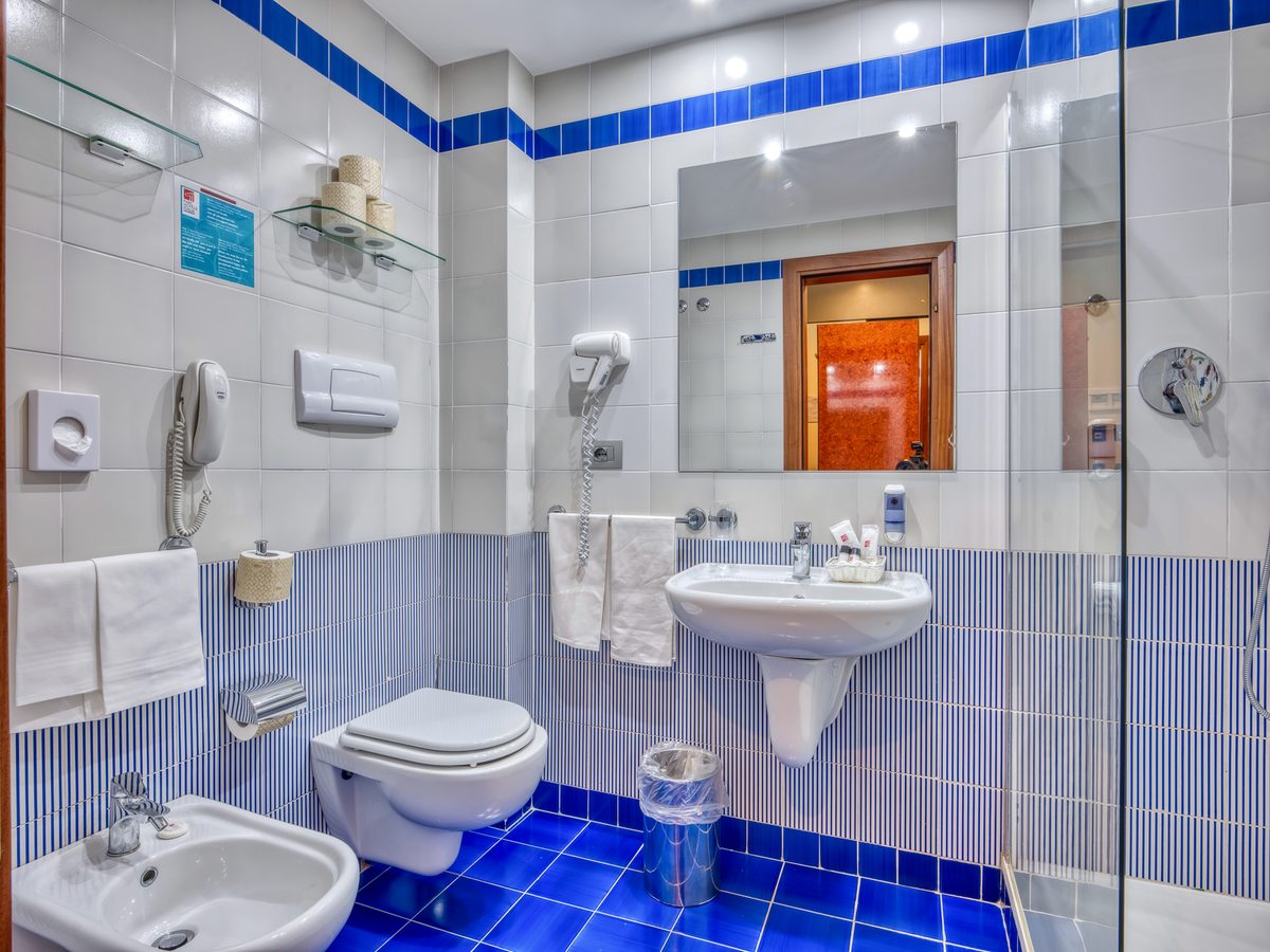 A hotel bathroom with blue and white tiles, a glass shower cubicle, a wall-mounted basin, a toilet and a bidet.