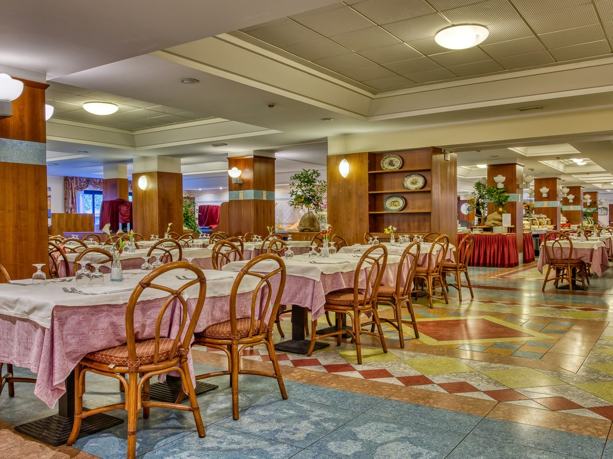 Interior of the main hotel restaurant with rattan chairs, tables laid with pink and white cloths, wooden pillars and a buffet counter along the back wall.