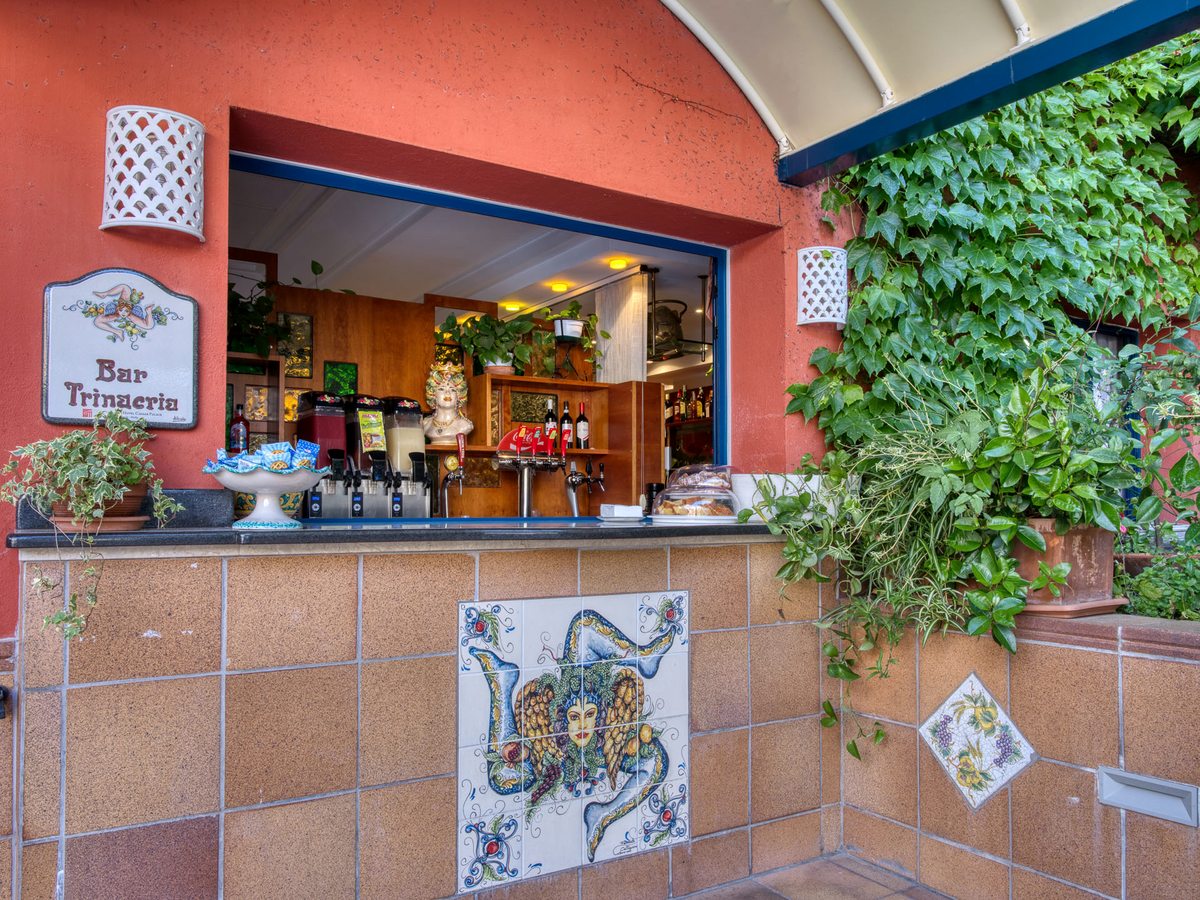 An outdoor service hatch in a terracotta-red wall, signed Bar Trinacria, its counter tiled with a Sicilian Trinacria emblem, drink dispensers and bottles inside and ivy alongside.