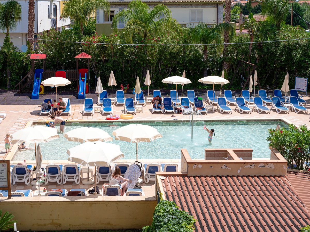 A shallow children's pool with a low water jet and parasols standing in the water, a small playground with slides beside it and rows of sun loungers around.