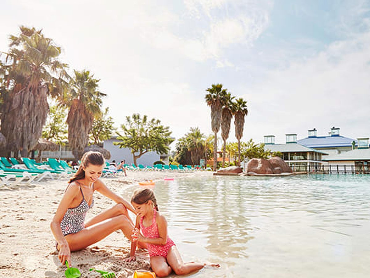 A woman and a young girl building sandcastles at the white-sand shallow edge of the pool, sunloungers and palms behind.