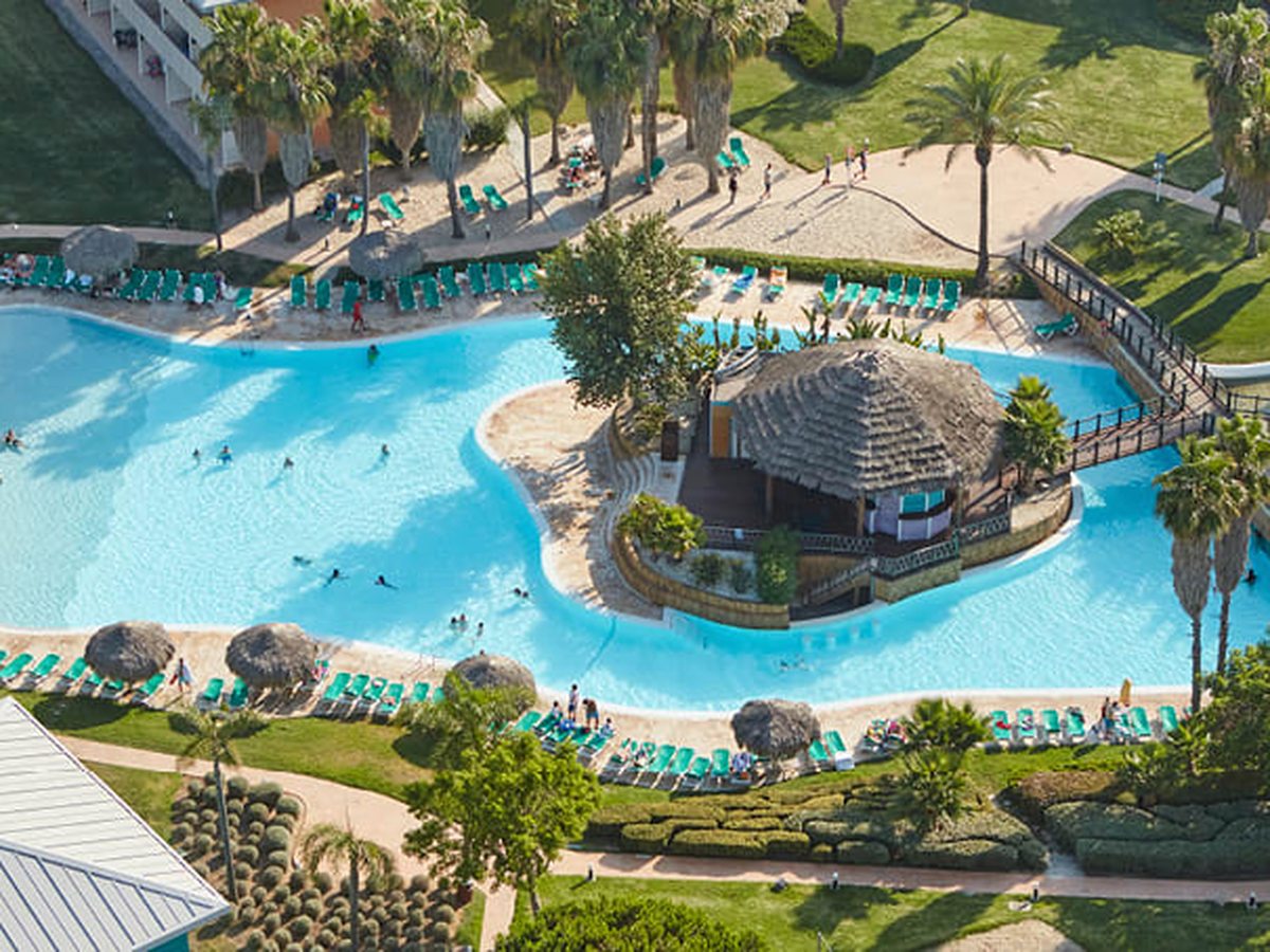An aerial view of the large lagoon-shaped pool with sandy shores, a thatched bar on a central island ringed by green sunloungers and gardens.