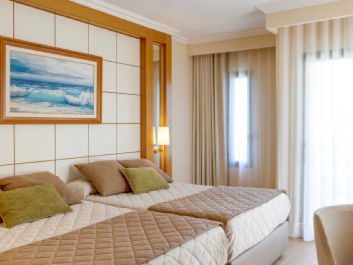 A Standard Room set up with two single beds pushed together beneath a framed seascape, next to a bright curtained window.