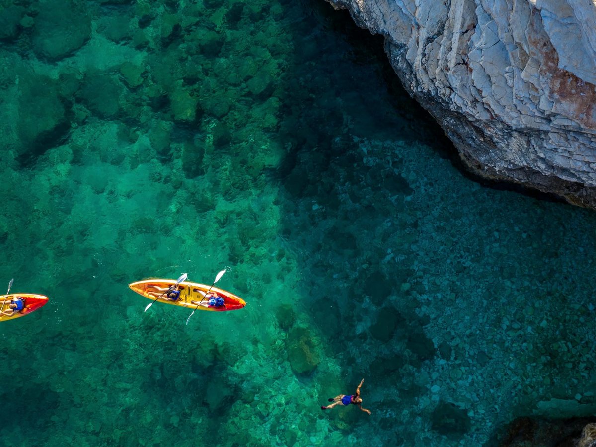 An aerial view of two kayakers and a swimmer in clear turquoise water beside pale limestone rocks.