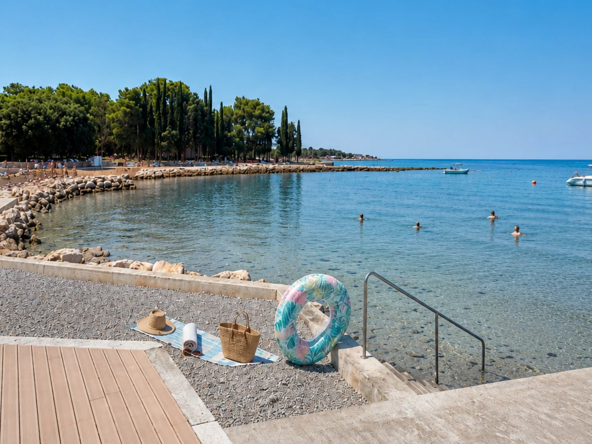 A swim ring and a bag beside steps leading into shallow clear water off a paved and pebble shore, with a pine-covered point beyond.
