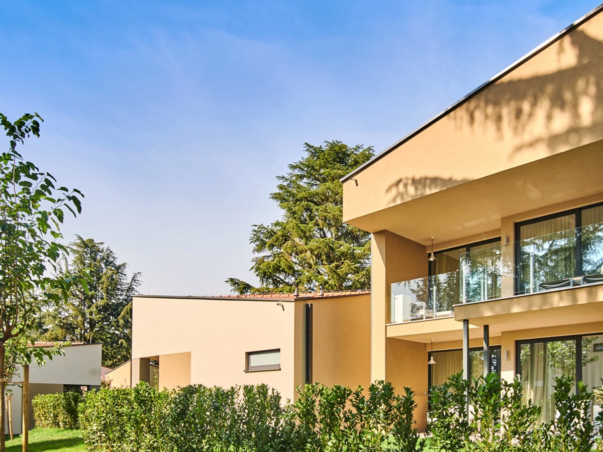 A tan two-storey guest building with recessed balconies and glass balustrades, a tall pine and a young tree beside it, and clipped hedges along the lawn under a clear blue sky.