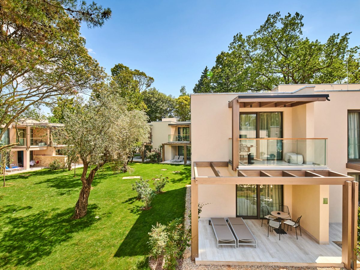 Cream two-storey garden rooms with terraces, loungers and glass balconies, set on lawns among olive and pine trees.