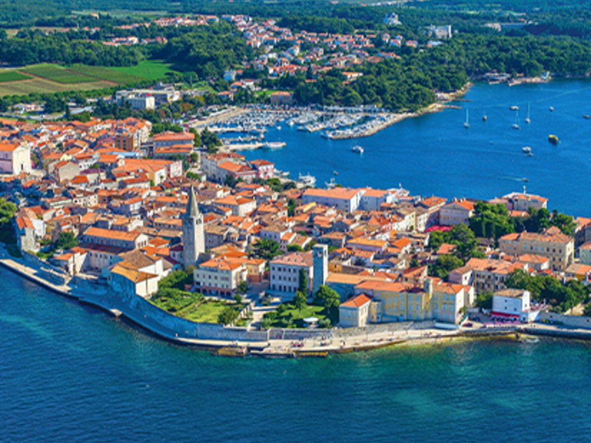An aerial view of a red-roofed old town on a peninsula with a marina and a basilica bell tower, ringed by clear blue sea.