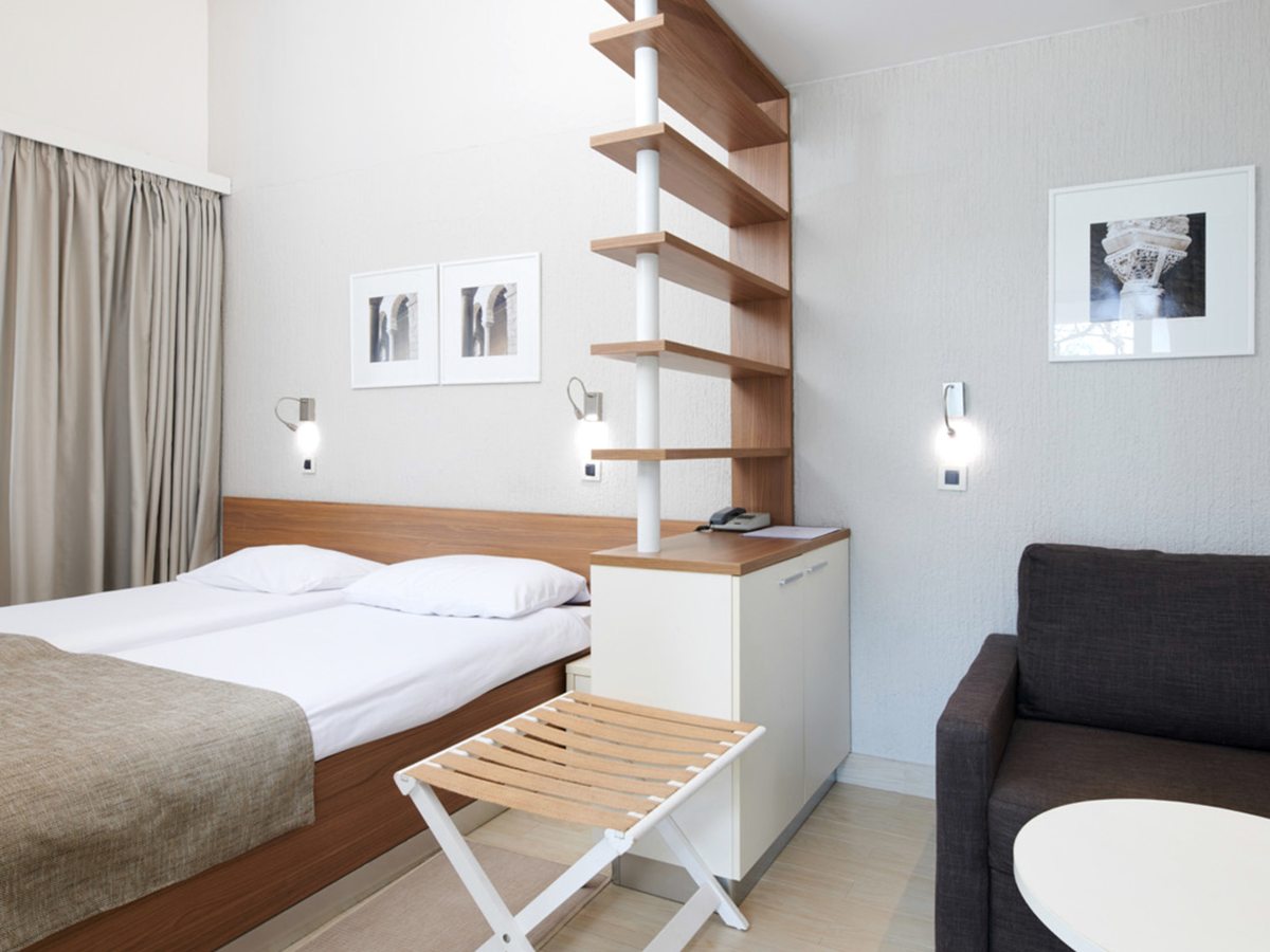 A room with two single beds together, a wooden headboard, a shelved divider, a folding luggage stool and a dark two-seat sofa.
