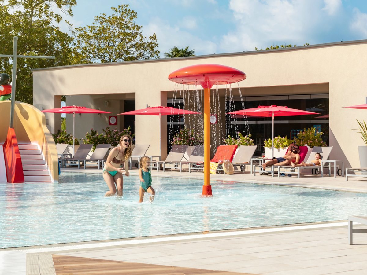 A woman and a toddler wade in a shallow pool under a red mushroom water sprayer, with a pirate-ship play feature, loungers and red parasols by a cream building.