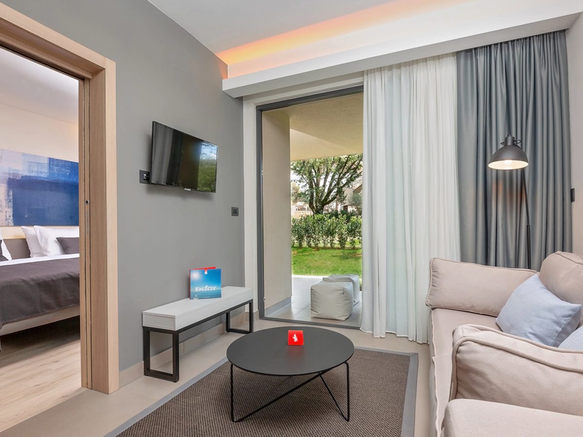 A suite living room with a beige sofa, a wall-mounted TV and a coffee table, the bedroom visible through an open doorway and a garden terrace beyond.