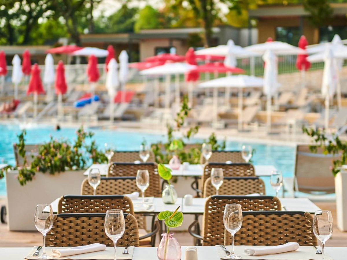Outdoor tables set with wine glasses and woven chairs on a deck beside a blue pool lined with red and white parasols.