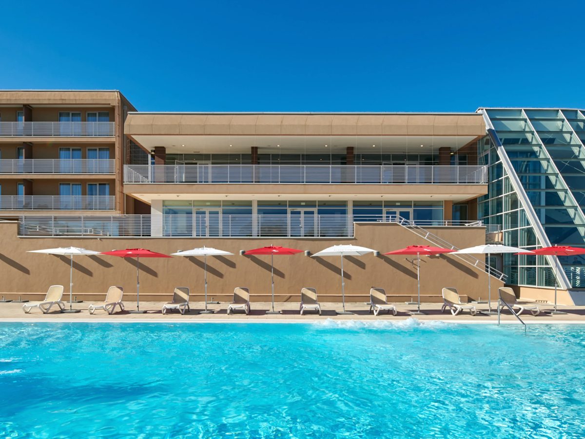 A large rectangular outdoor pool with red and white parasols and loungers along a wall, beside a cream building with a triangular glass atrium.