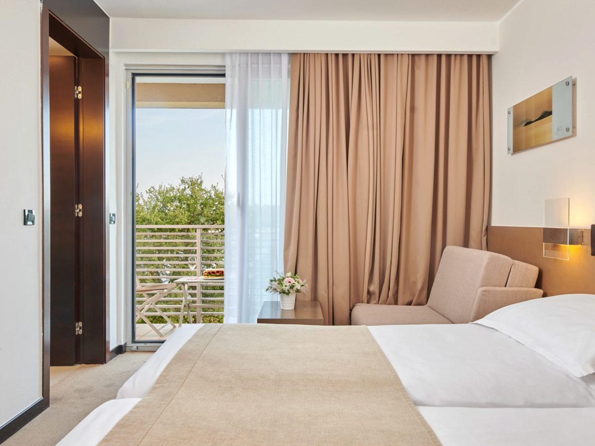 A room with a made-up bed, a beige runner and a small sofa, and a balcony with a table and chairs opening onto greenery.