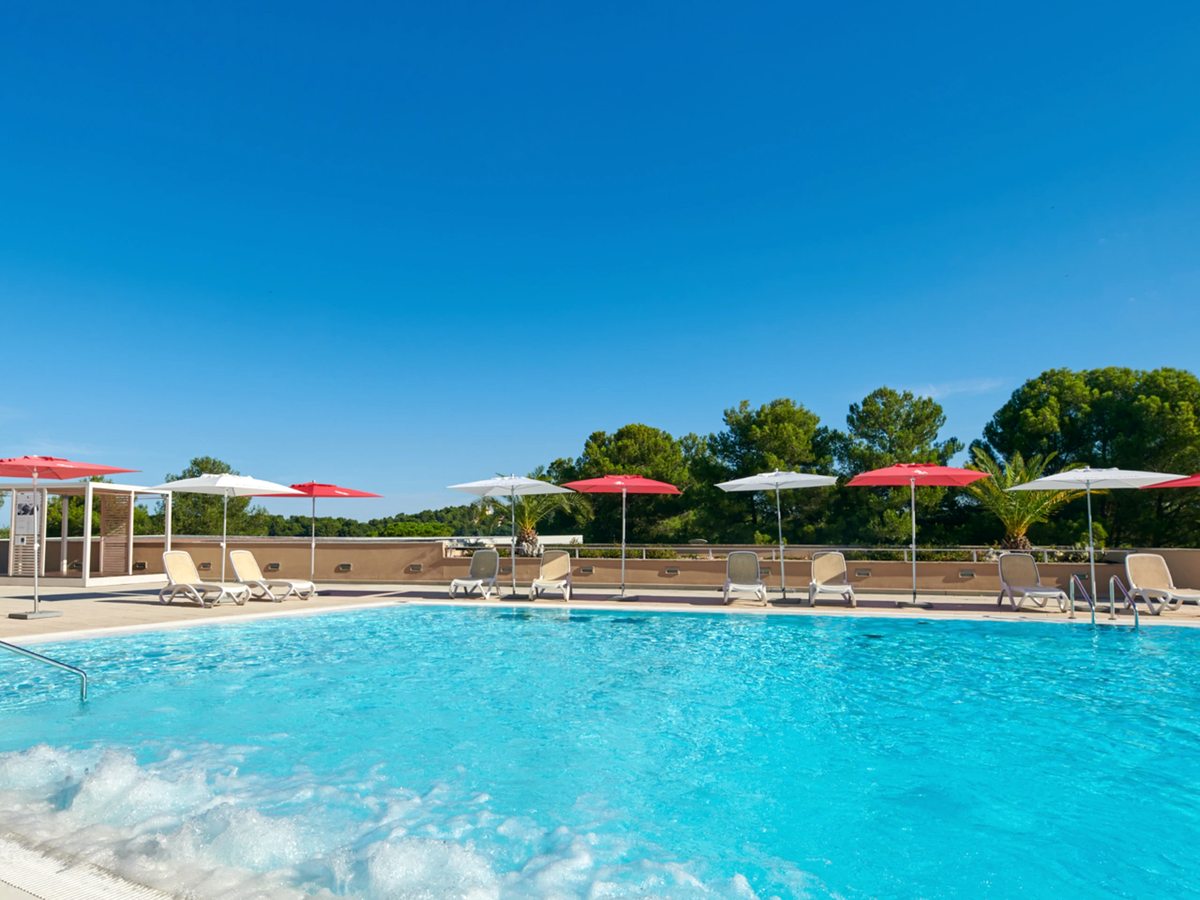 A large freeform outdoor pool with red and white parasols and white loungers, backed by tall pine trees under a clear blue sky.