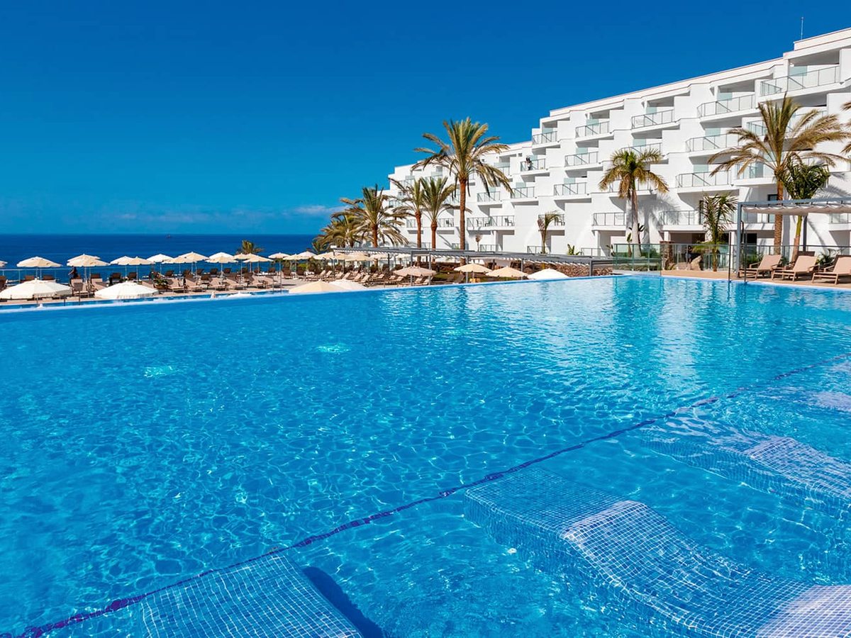 A large freshwater pool edged with loungers and parasols, the white guest-room tower and palms behind, sea to the left.