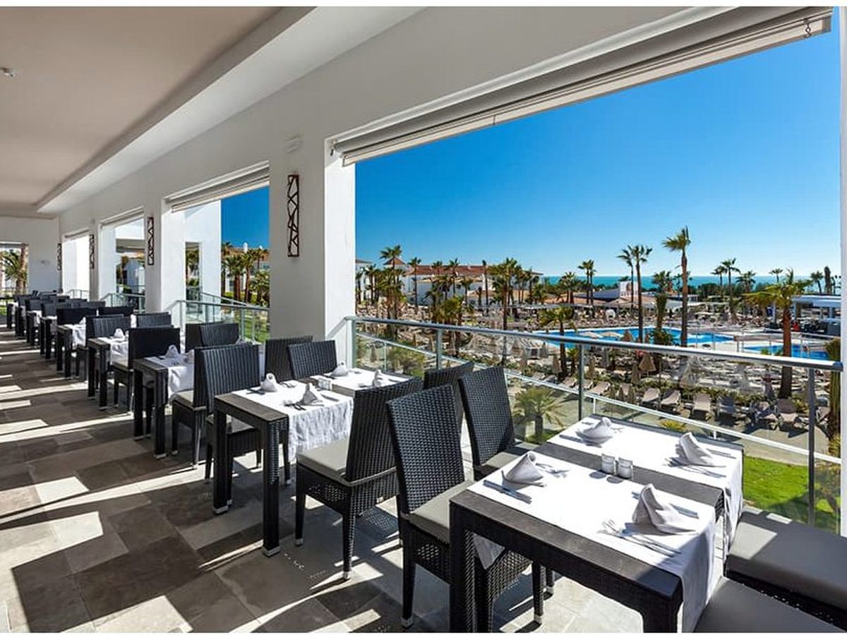 A covered restaurant terrace with white-linen tables and black chairs, looking out over the pools, palms and sea.