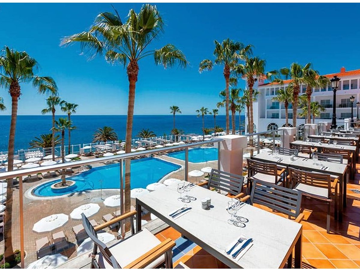 Restaurant terrace with laid tables and terracotta floor tiles, looking past palms and the outdoor pool to the Atlantic.