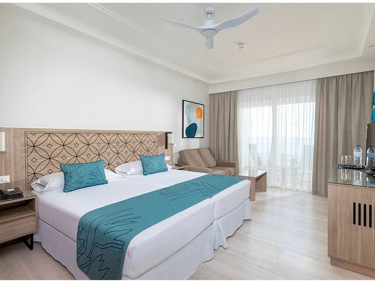 Guest room with two beds pushed together under a patterned headboard, a sofa beside the balcony doors and a TV on a wooden unit.
