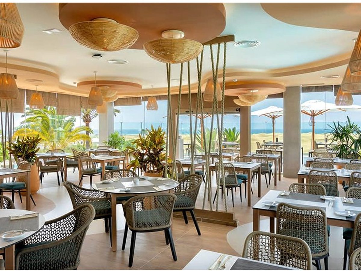 Bright buffet restaurant with woven chairs, rattan pendant lights and full-height windows onto palms and the sea.