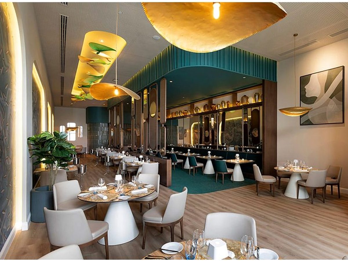 À la carte dining room in teal and gold, with cushioned chairs, patterned walls and gold pendant lights over a wood floor.
