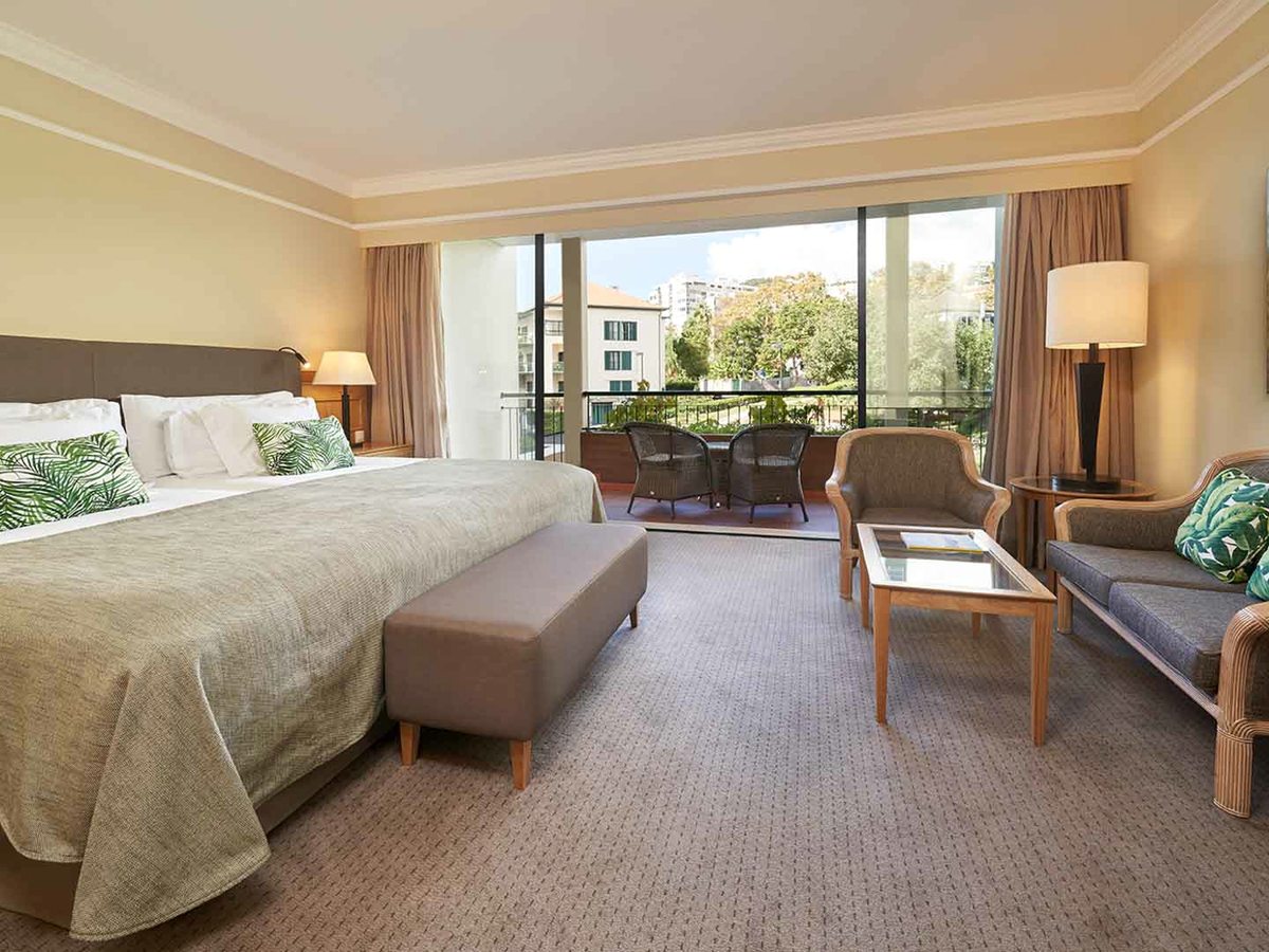 A room with a king bed, a sofa and armchairs around a low table, and sliding doors to a furnished balcony looking over greenery.