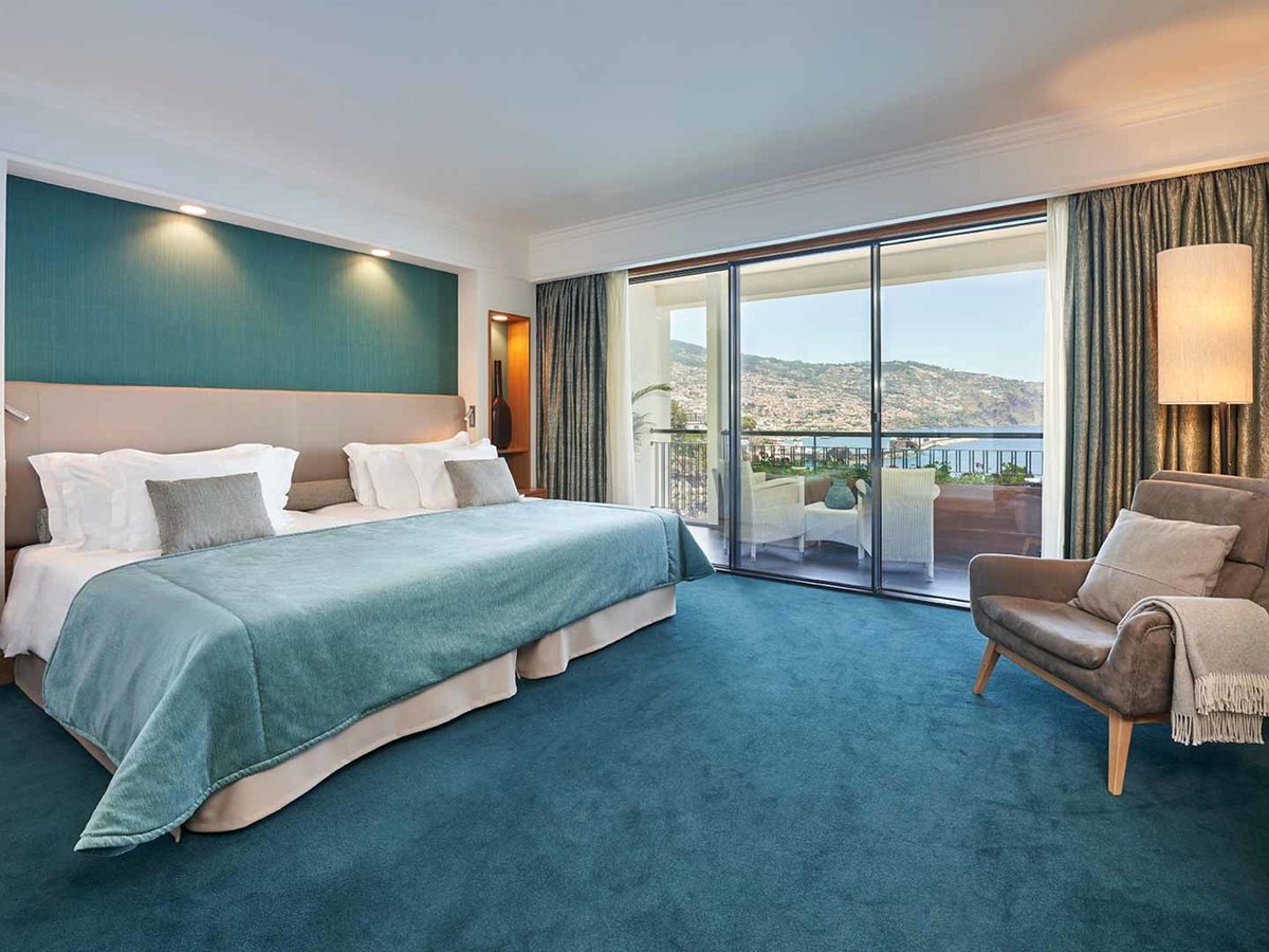 A room with a teal feature wall and carpet, a king bed and an armchair, sliding doors to a balcony overlooking Funchal's hillside and bay.