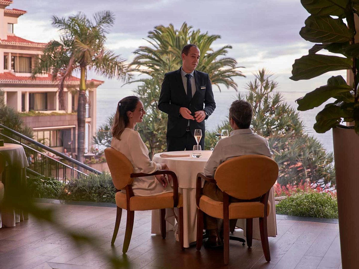 A waiter serving a couple at a table laid with wine on a garden terrace, palms and the hotel facade behind.