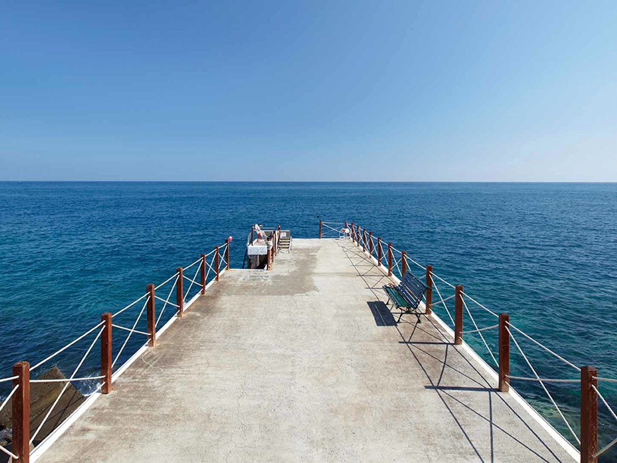 A concrete pontoon with rope railings reaching out over clear blue water toward the open sea.