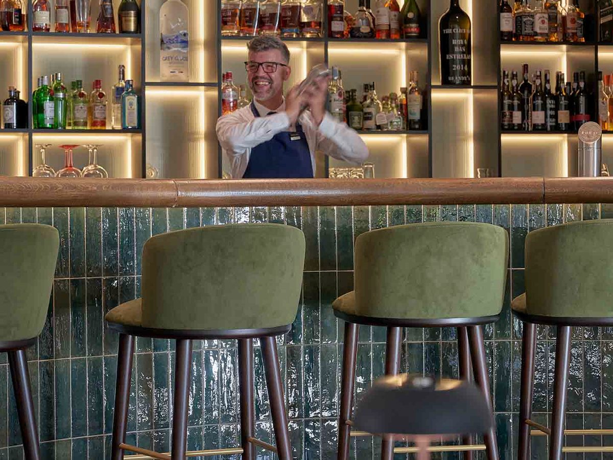 A bartender in a Cliff Bay apron shaking a cocktail behind a green-tiled bar counter lined with green velvet stools and backlit spirit shelves.