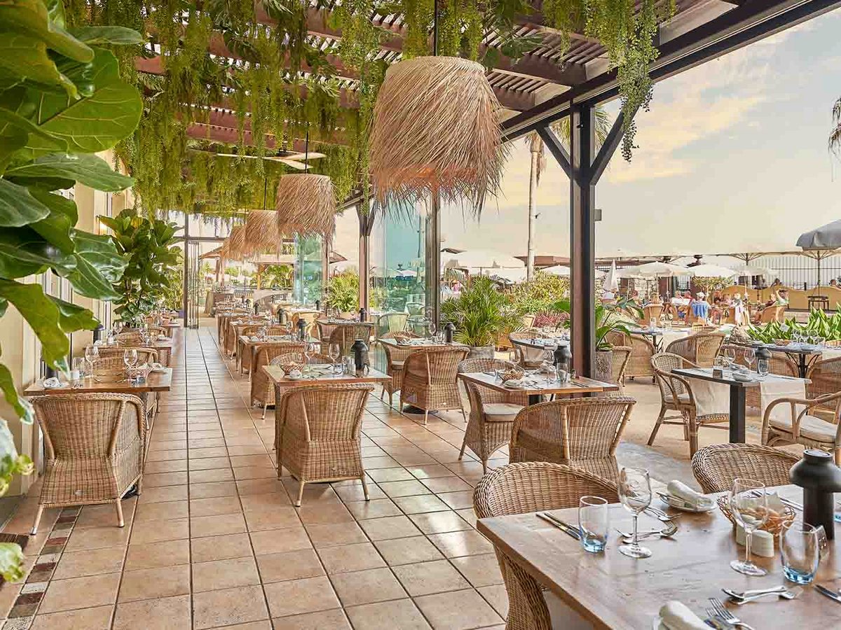 A covered open-air restaurant terrace with wicker armchairs, thatched pendant lights and hanging greenery, opening to the pool and sea beyond.