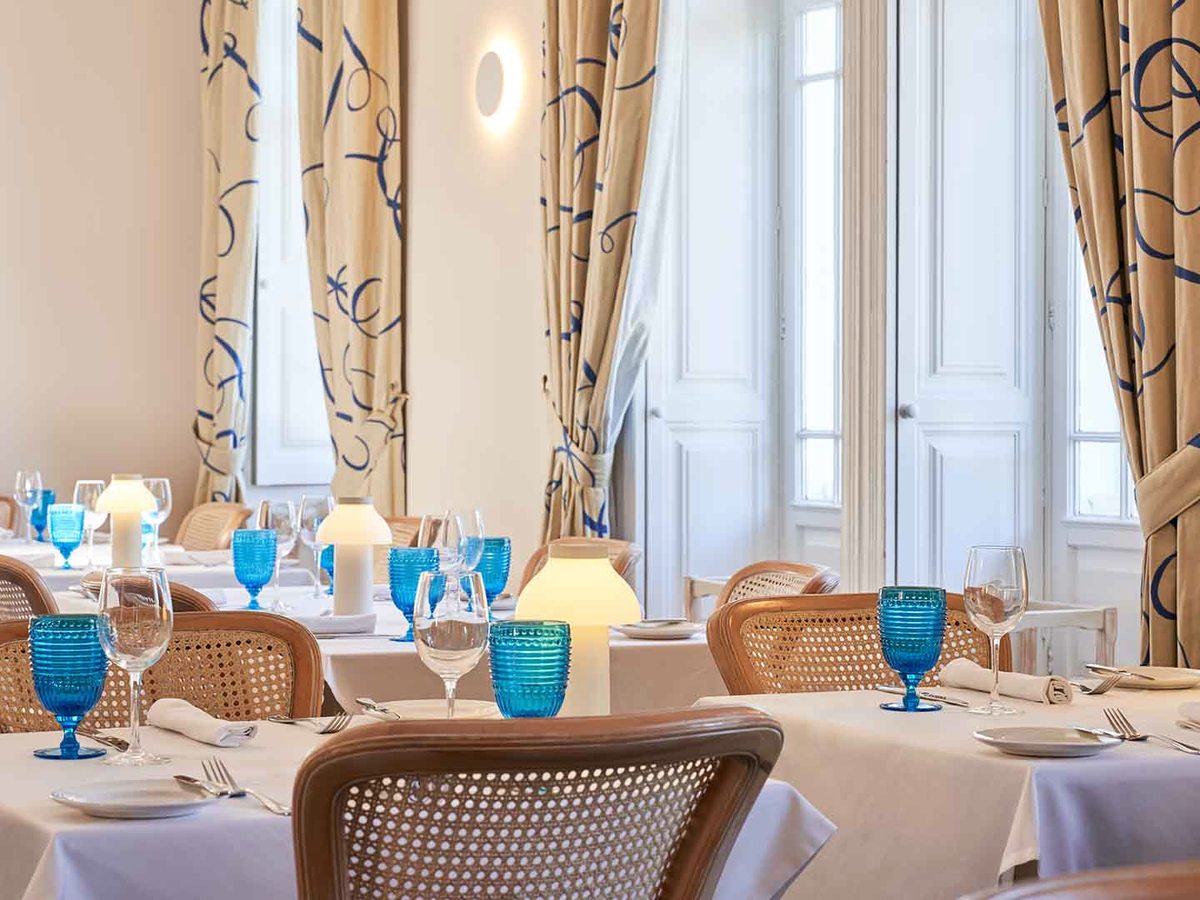 An indoor restaurant with cane chairs, white-clothed tables, blue glassware and tall curtained windows.