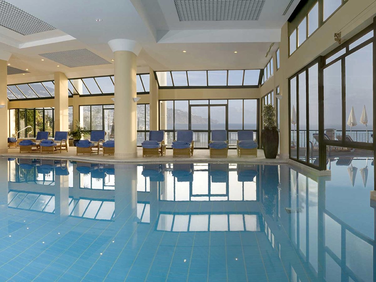 A glass-roofed indoor pool with blue loungers along the tiled surround and floor-to-ceiling windows looking out to the sea.