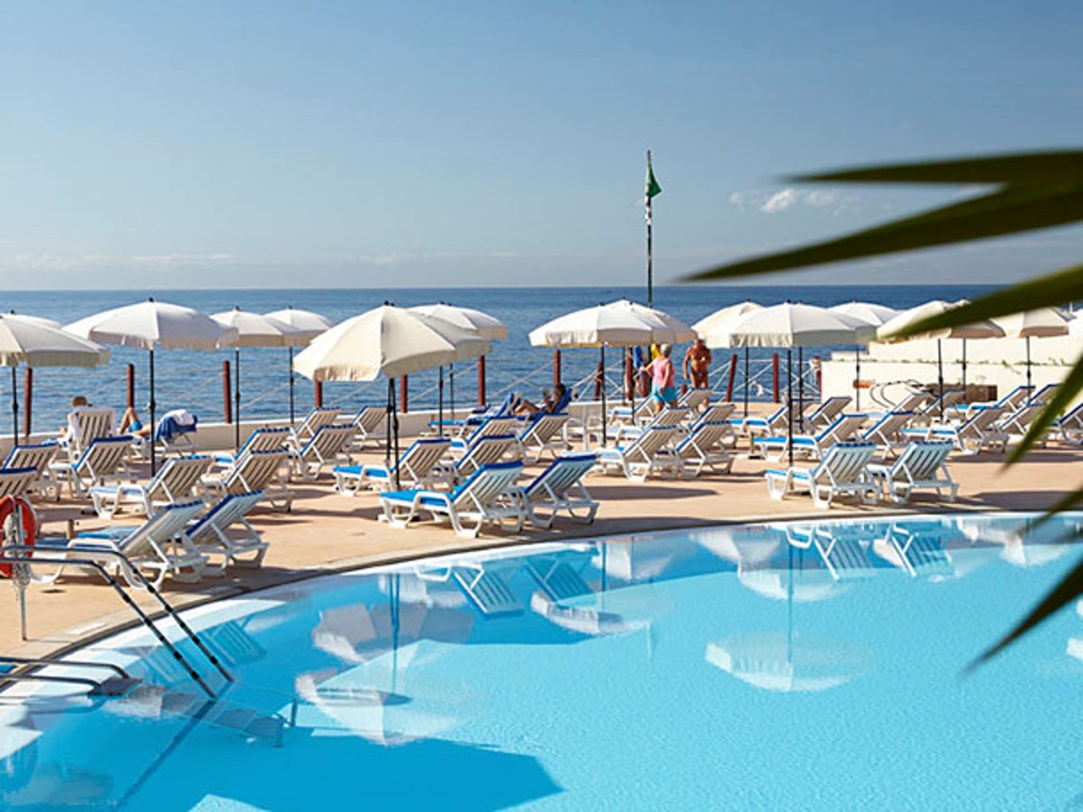 A curved outdoor pool at sea level edged with blue-and-white loungers and white parasols, the Atlantic directly beyond a green safety flag.