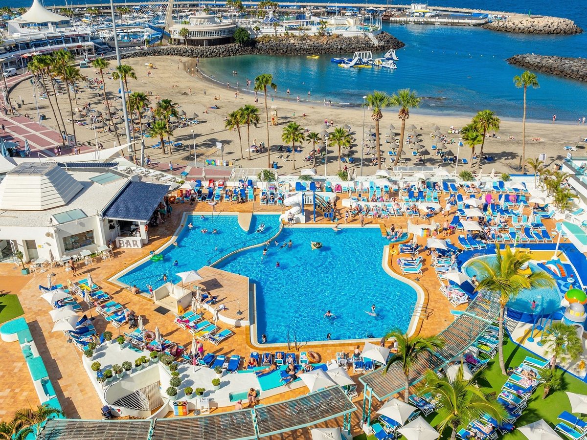 An aerial view of the curved main pool ringed by loungers, with the beach and marina behind.