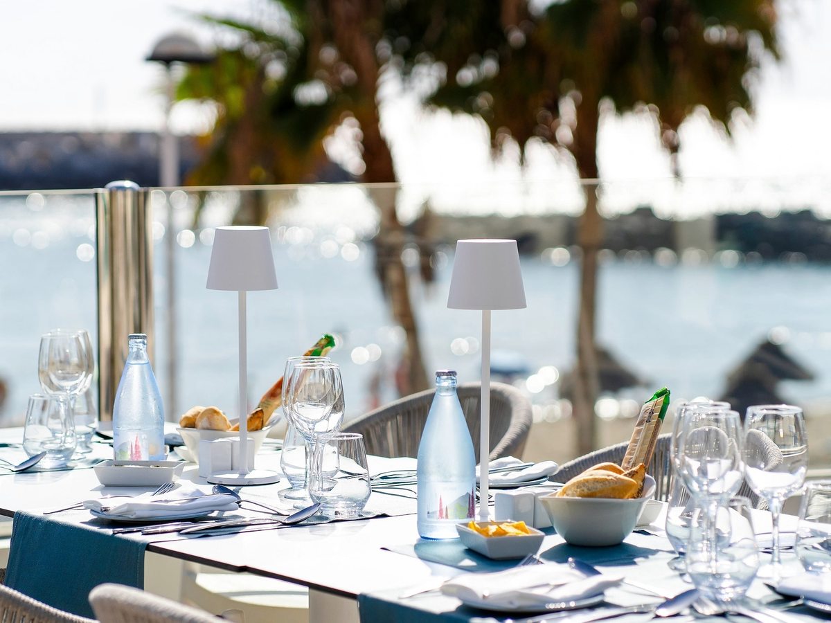 An outdoor restaurant table laid with glasses and bread on a terrace among palms by the marina.