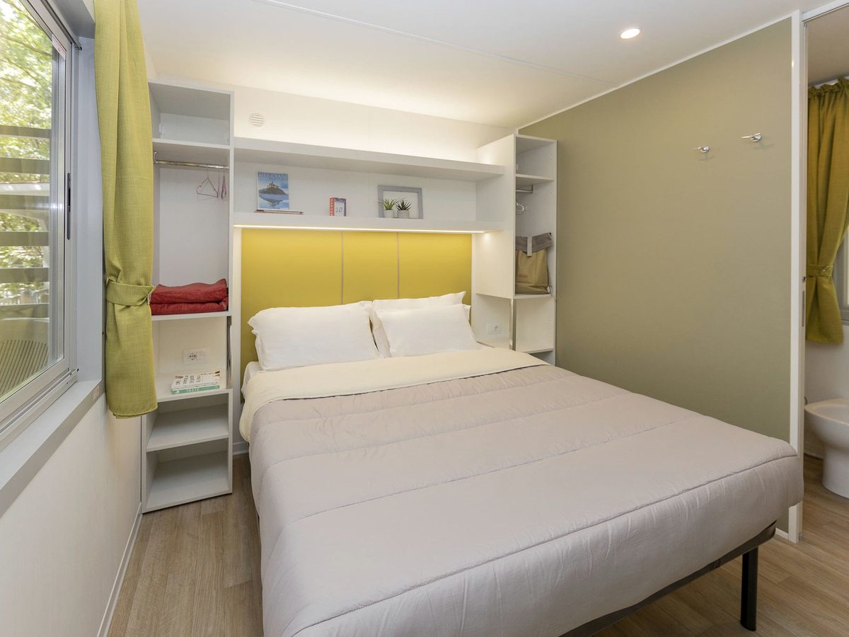 A double bed with a padded yellow headboard and white shelving in a bright bedroom.