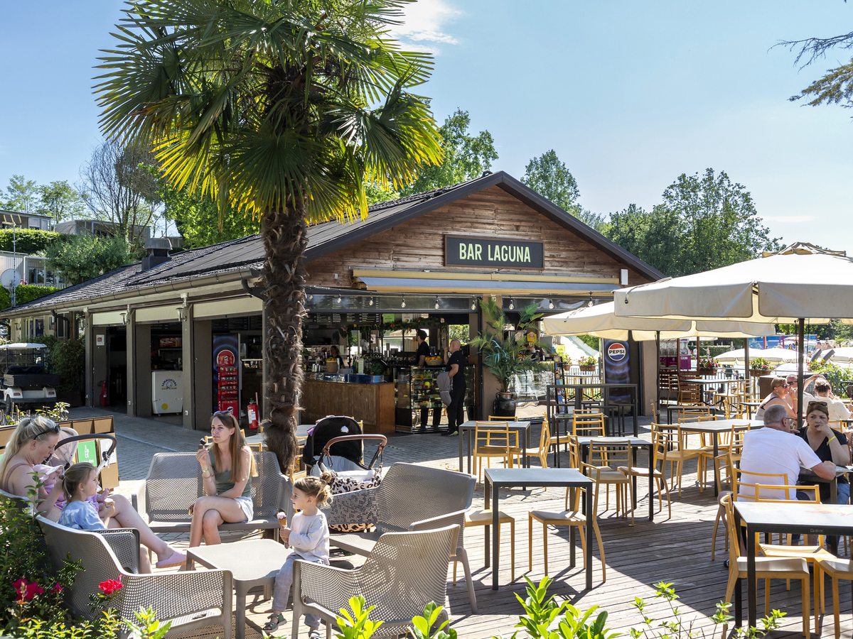 An open-sided bar signed Bar Laguna, with parasols, a palm tree and outdoor tables.