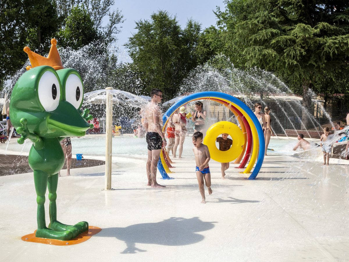 A splash area with arched water jets and a large green frog mascot, children running through the spray.