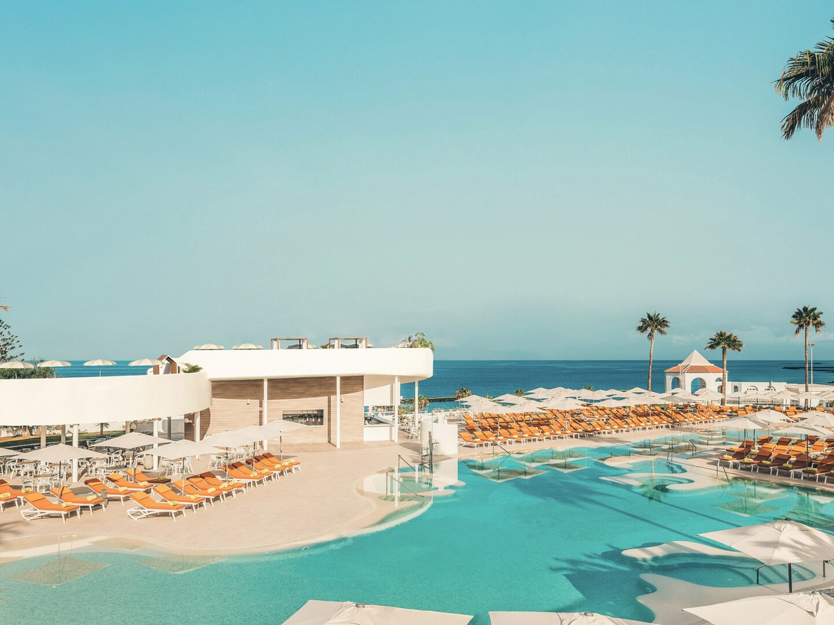 A pool on a white terrace with orange sun loungers and parasols, palms and the sea beyond.
