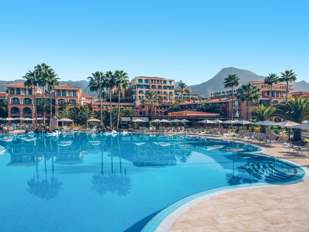 A large curved pool edged with sun loungers, the terracotta hotel blocks and palms behind and a mountain in the distance.
