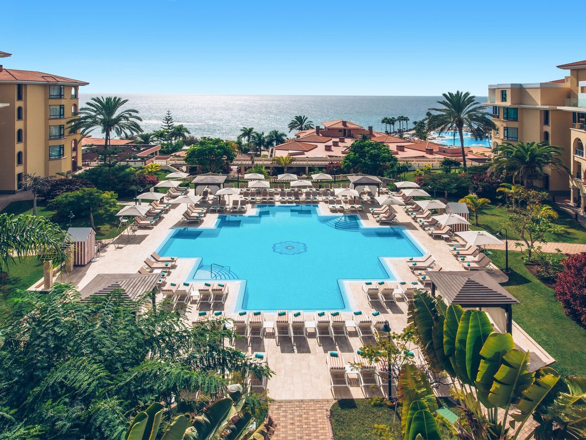Aerial view of the resort's freeform and rectangular pools among lawns and palms, with the hotel blocks and the sea beyond.