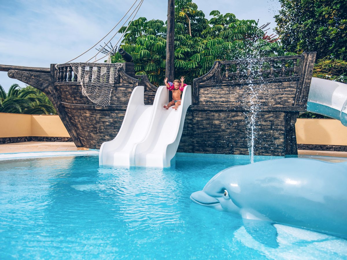 A shallow children's pool with a pirate-ship play structure, twin white water slides and a dolphin water feature.