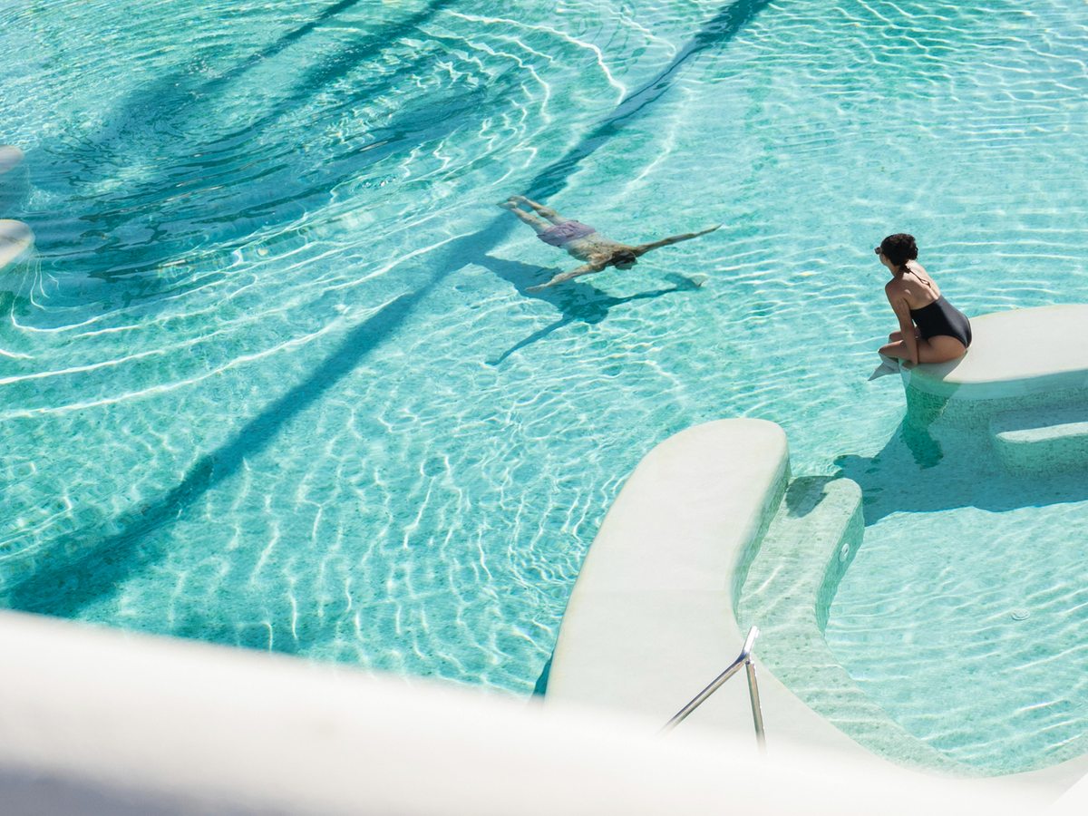 A turquoise pool with built-in in-water loungers along a shallow ledge and guests swimming, with steps at one side.