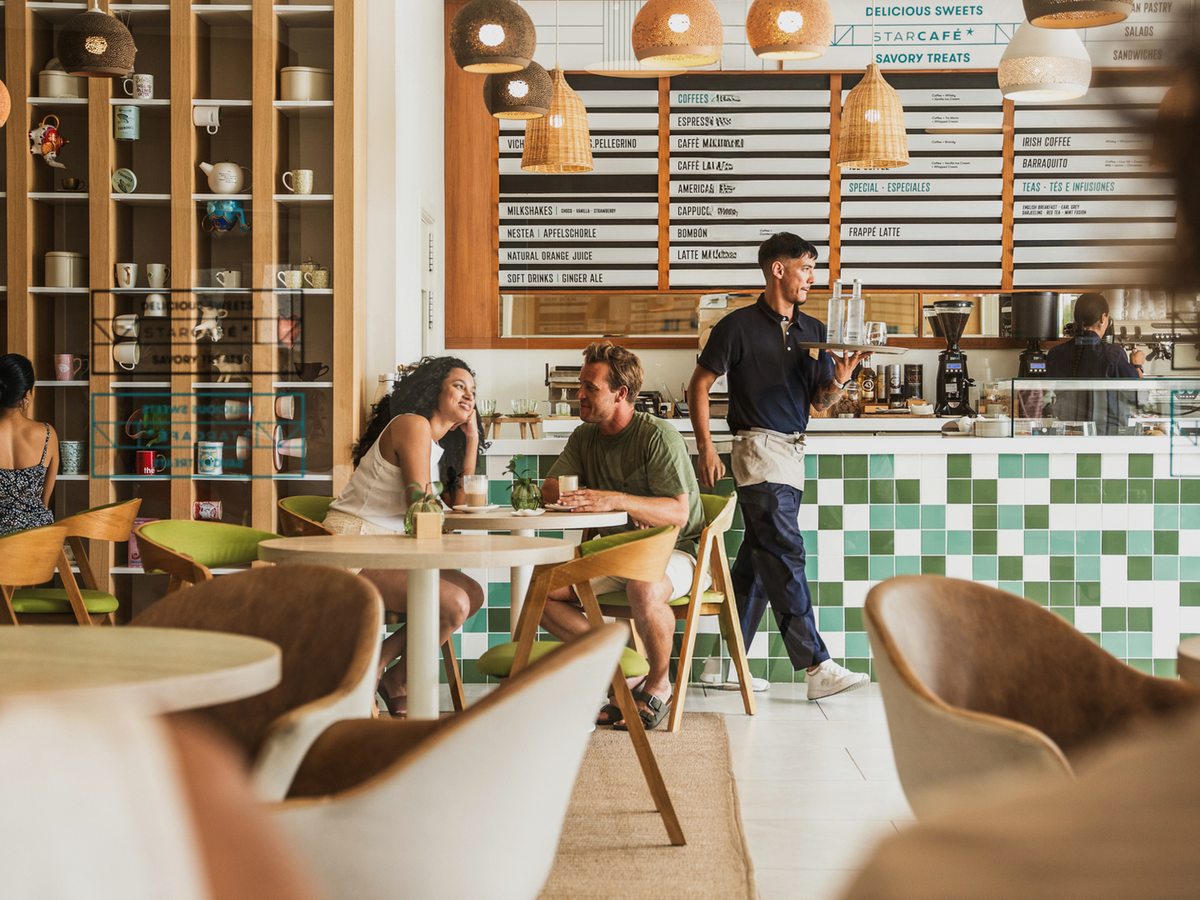 An indoor café-bar with a green-tiled counter, a chalk menu board, low tables and cane chairs, with staff serving guests.